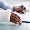 Overhead View Of Injured Man With Bandage Hand Filling Insurance Claim Form On Clipboard