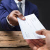 Close-up Of A Businessman's Hand Giving Cheque To Colleague Over Wooden Desk